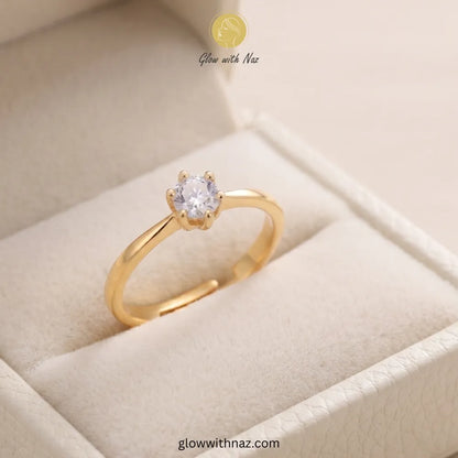 A gold ring with a single round clear gemstone in a prong setting, displayed inside a cream-colored jewelry box. The brand name 'Glow with Naz' is visible at the top, and a website URL is visible at the bottom.