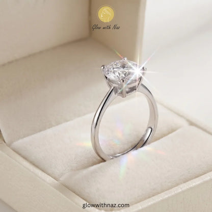 A silver solitaire engagement ring with a round brilliant-cut diamond, displayed inside a cream-colored velvet ring box. The image features a stylized starburst and rainbow light flare effect around the diamond, with the brand name 'Glow with Naz' visible in the upper portion.