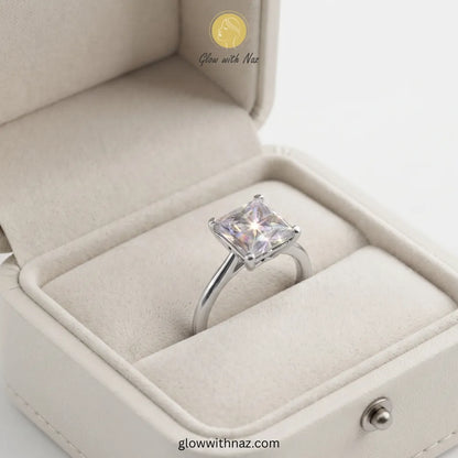 A square princess-cut solitaire ring with a silver-colored band displayed inside an open cream-colored velvet ring box. The ring features a sparkling clear gemstone.