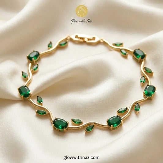 A gold leaf-shaped bracelet adorned with green marquise and oval gemstones, displayed on a draped cream satin fabric. The brand logo 'Glow with Naz' is visible at the top. Tulip Rapunzel bracelet, floral tulip bracelet, Rapunzel inspired jewelry, aesthetic tulip bracelet, trendy charm bracelet, stainless steel tulip bracelet, gold plated flower bracelet, delicate floral wrist accessory, cute aesthetic jewelry, viral tulip bracelet, Rapunzel theme accessories, dainty floral bracelet, crystal tulip charm, sta
