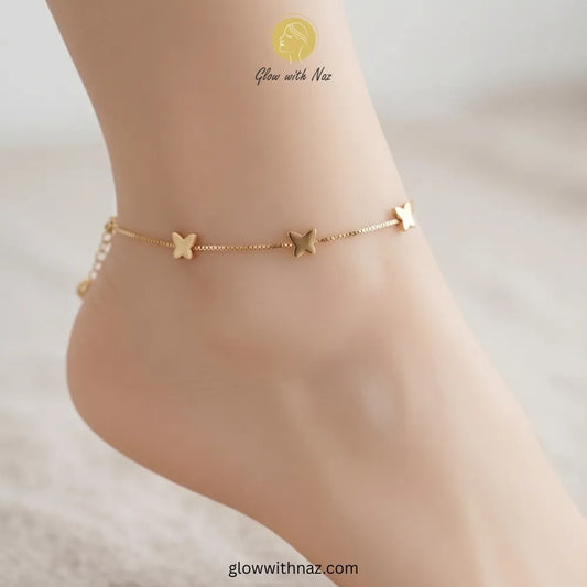 Flat lay of a delicate gold butterfly anklet featuring a sturdy box chain and a small bell charm by Glow With Naz