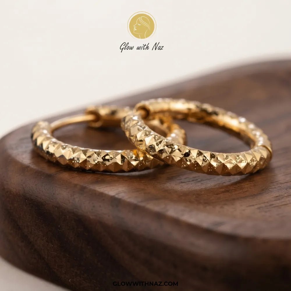Gold Textured Diamond-Cut Bali Hoop Earrings - #Gold Textured Diamond-Cut Bali Hoop EarringsJewelryGlow With NazGlow With NazE089A pair of textured gold hoop earrings resting on a dark wooden block, with a 'Glow with Naz' brand logo at the top and a website URL at the bottom.#