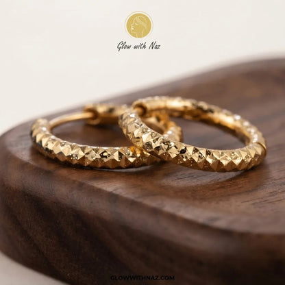 Gold Textured Diamond-Cut Bali Hoop Earrings - #Gold Textured Diamond-Cut Bali Hoop EarringsJewelryGlow With NazGlow With NazE089A pair of textured gold hoop earrings resting on a dark wooden block, with a 'Glow with Naz' brand logo at the top and a website URL at the bottom.#