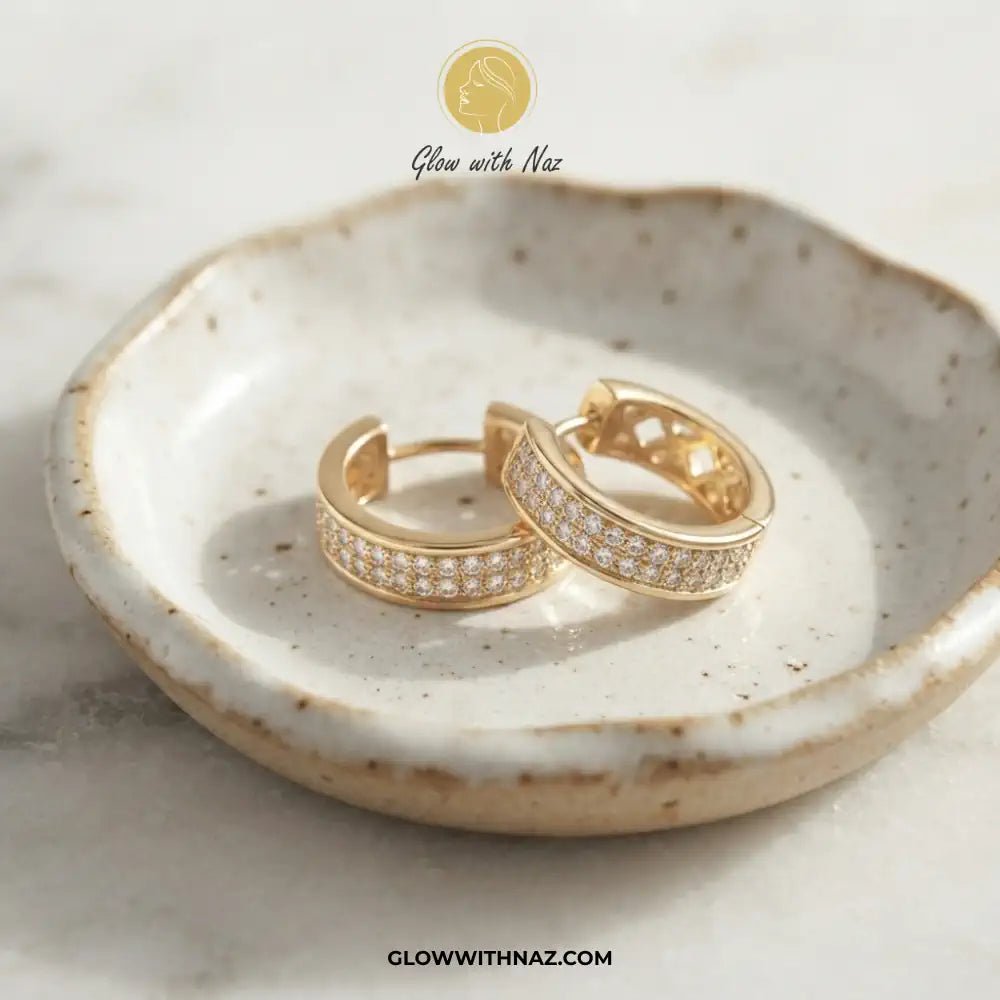 Golden Zircon Mid Size Slide Hoop for Women - Golden Zircon Mid Size Slide Hoop for WomenJewelryGlow With NazGlow With NazE058A pair of gold huggie hoop earrings with pavé-set clear stones, displayed on a speckled ceramic dish. The image features a 'Glow with Naz' logo at the top and a website URL at the bottom.