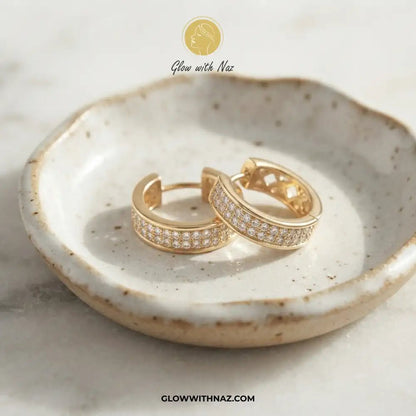 Golden Zircon Mid Size Slide Hoop for Women - Golden Zircon Mid Size Slide Hoop for WomenJewelryGlow With NazGlow With NazE058A pair of gold huggie hoop earrings with pavé-set clear stones, displayed on a speckled ceramic dish. The image features a 'Glow with Naz' logo at the top and a website URL at the bottom.