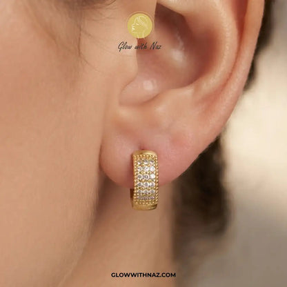 Minimal Zircon Mini Thick Hoops Earring for Girls - Minimal Zircon Mini Thick Hoops Earring for GirlsJewelryGlow With NazGlow With NazE055A close-up of a person's ear wearing a small gold huggie hoop earring set with clear pavé stones. The image includes a watermark logo for 'Glow with Naz' and the website 'GLOWWITHNAZ.