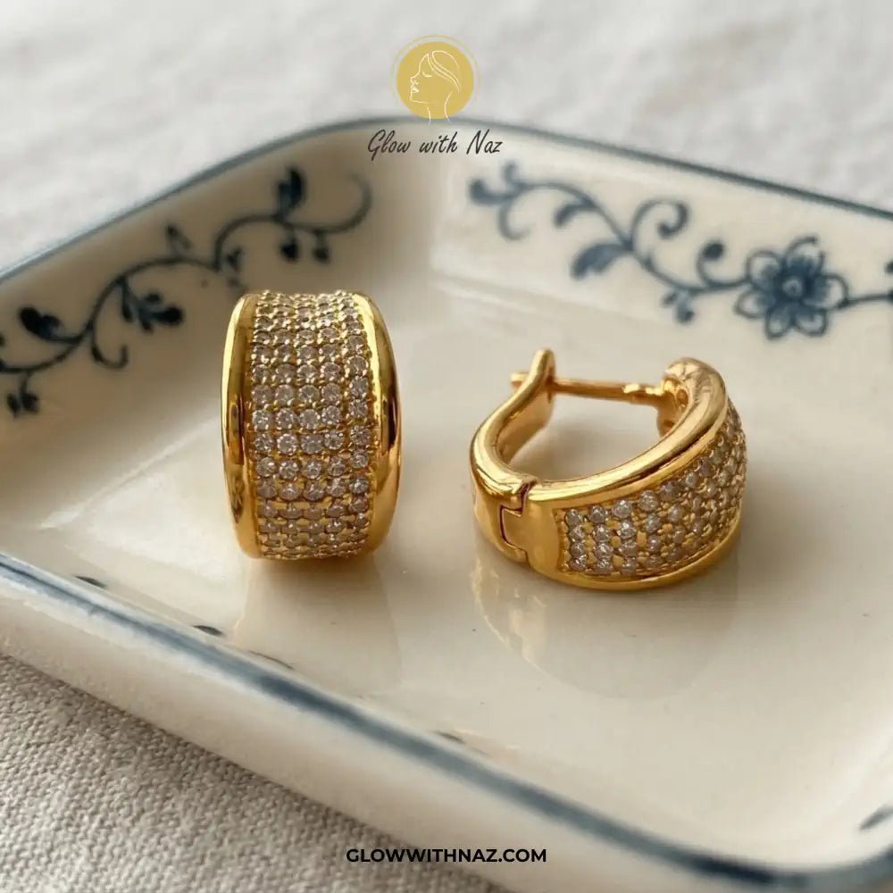 Minimalist Diamond Curvy Hoop Earrings for Girls - Minimalist Diamond Curvy Hoop Earrings for GirlsJewelryGlow With NazGlow With NazE056A pair of gold hoop earrings with pavé-set clear stones, displayed on a white ceramic dish with blue floral detailing. The brand logo 'Glow with Naz' is visible at the top, and the URL 'GLOWWITHNAZ.