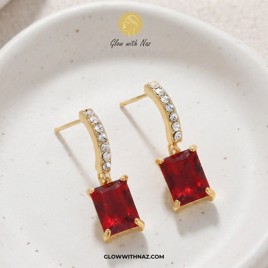 Premium Square Drop Zircon Stud Earring for Girls - Premium Square Drop Zircon Stud Earring for GirlsJewelryGlow With NazGlow With NazE035A pair of gold drop earrings with rectangular red gemstones and a curved top set with small clear stones, displayed on a white speckled ceramic dish. The brand name 'Glow with Naz' is visible at the top of the image. studs earrings, small earrings, girls earrings, simple earrings, earrings for girls, black earrings, earrings, ear rings, earrings tops, stud ear