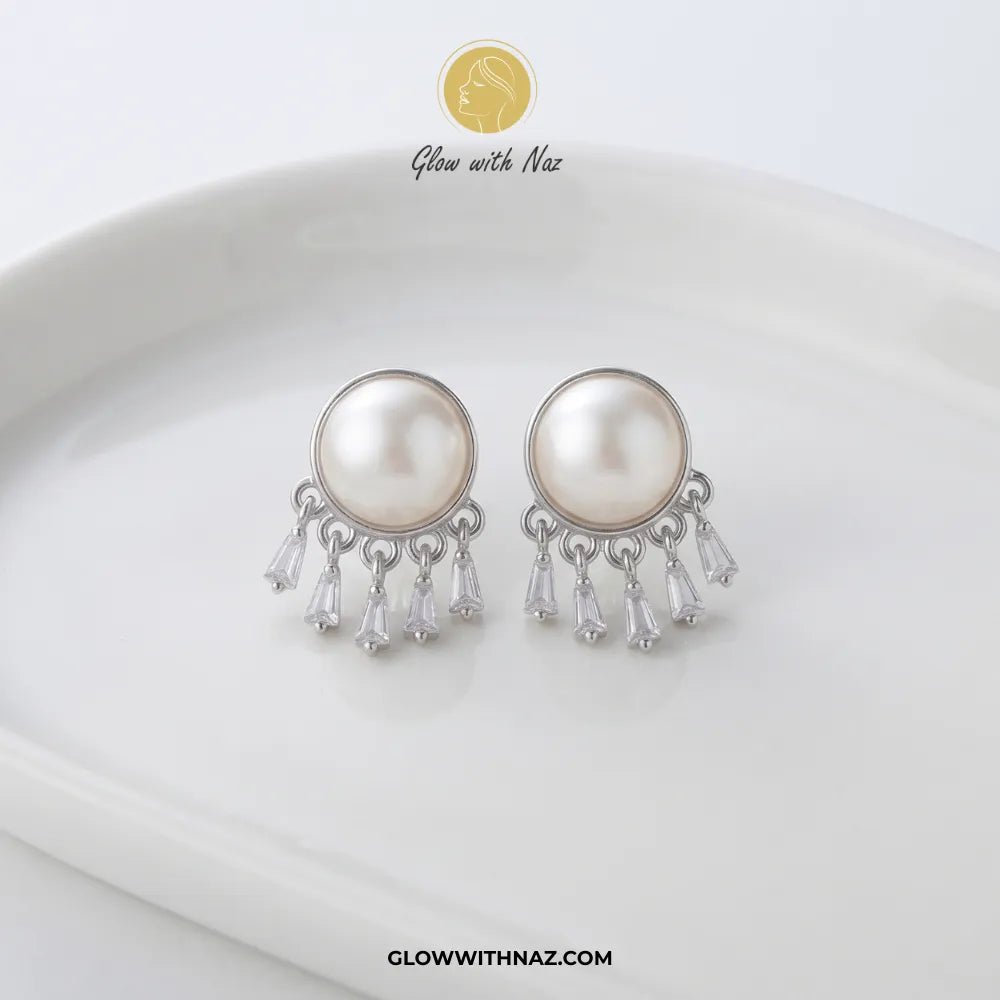 Radiant Mini Silver Pearl Stud Earring - #Radiant Mini Silver Pearl Stud EarringJewelryGlow With NazGlow With NazE087A pair of silver-toned dangle earrings with large round white faux pearls and clear teardrop crystal drops, displayed on a white ceramic dish. The brand logo 'Glow with Naz' is visible at the top and the website 'GLOWWITHNAZ.#