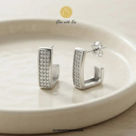 Silver Zircon Square Statement Earring for Women - Silver Zircon Square Statement Earring for WomenJewelryGlow With NazGlow With NazE054A pair of silver-toned square-shaped hoop earrings with pavé-set clear stones on one side, displayed on a round cream-colored ceramic plate. The brand logo 'Glow with Naz' is visible at the top, and the website 'GLOWWITHNAZ.