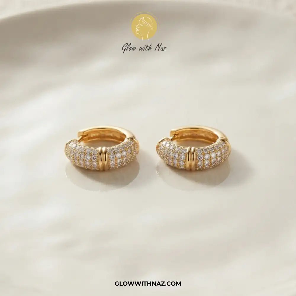 Zircon 3 Bar Mini Hoops Earrings - #Zircon 3 Bar Mini Hoops EarringsJewelryGlow With NazGlow With NazE057A pair of gold huggie hoop earrings paved with small clear stones, displayed on a soft cream-colored surface. The brand logo 'Glow with Naz' is visible at the top, and the website 'GLOWWITHNAZ.#