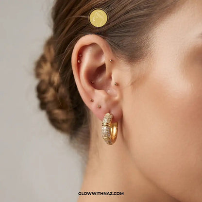 Zircon 3 Bar Mini Hoops Earrings - #Zircon 3 Bar Mini Hoops EarringsJewelryGlow With NazGlow With NazE057Close-up of a model's ear showcasing a textured gold huggie hoop earring, with other gold ear piercings on the lobe and cartilage. A gold coin is placed on the model's head for scale, demonstrating the earring's size.#
